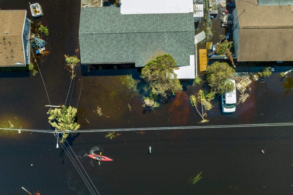 An aerial drone view of a flooded residential street with dark water surrounding houses and a person navigating a red kayak past submerged power lines.