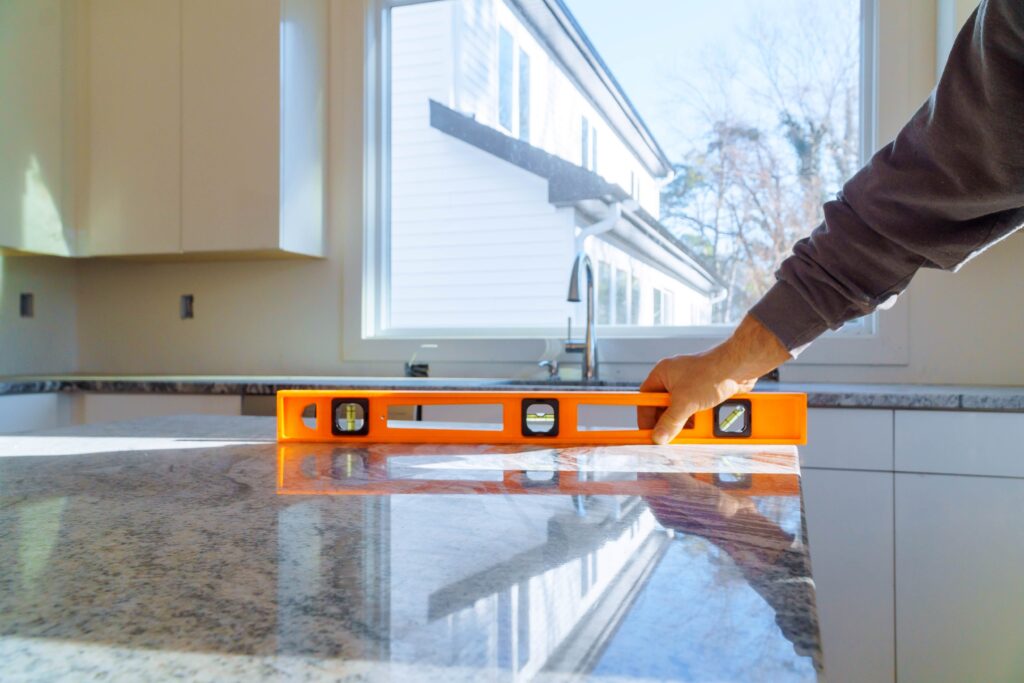 Close-up of a contractor's hand using an orange spirit level on a polished granite kitchen island countertop during installation, with a bright window and white kitchen cabinetry in the background.