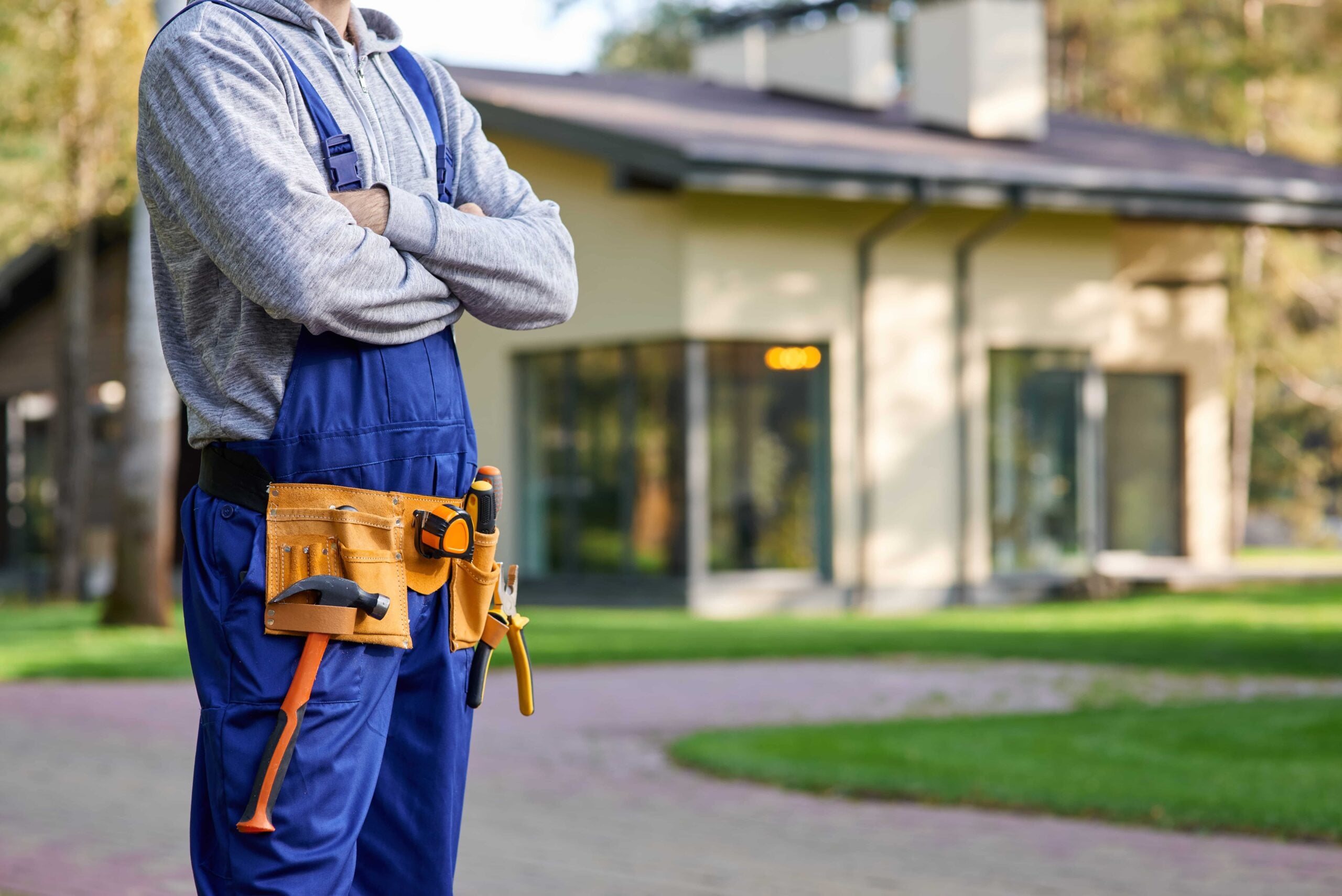 Preventative Home Maintenance to Protect Your Florida Property 1 A cropped shot of a professional home renovation contractor with crossed arms wearing blue overalls and a tool belt, standing in front of a modern suburban house in Tampa, Florida.